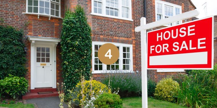 Brick house with a red House for Sale sign in front, surrounded by greenery and flowers.