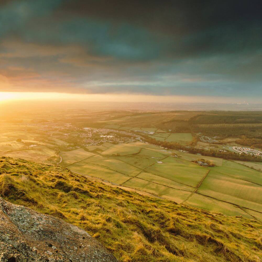 Sunset view over lush green fields from a hilltop, with a dramatic sky above.